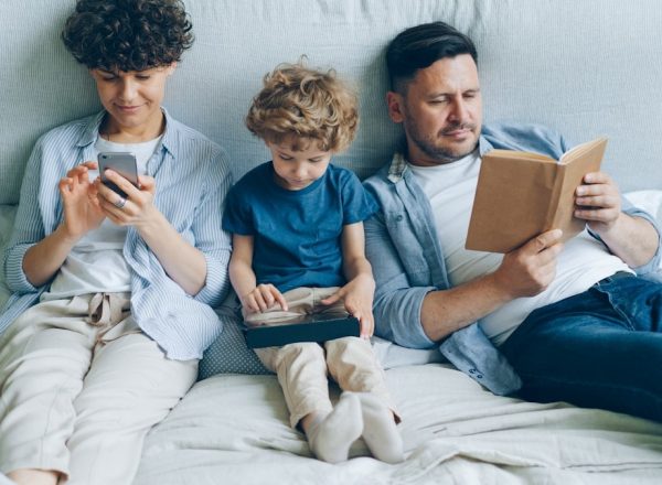 Mother using smartphone son holding tablet father reading book in bed at home